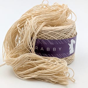 Può includere: Un gomitolo di filato color crema con un'estremità sciolta e srotolata. Una fascia di carta viola avvolge il centro del gomitolo, riportando il testo bianco "RABBY" e una sagoma bianca di un coniglio in piedi sulle zampe posteriori.