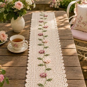 Könnte beinhalten: Ein rechteckiger, cremefarbener Häkel-Tischläufer mit einem zentralen Rankenmuster aus kleinen rosa und dunkelrosa Blüten. Der Läufer hat einen gewellten Rand und eine strukturierte Strickoberfläche, die sich vertikal über die Mitte erstreckt.