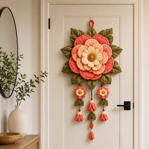 Peut inclure: Une décoration murale en tissu matelassé représentant une grande fleur centrale aux pétales superposés dans des tons corail, pêche et crème, entourée de feuilles vert foncé. Trois pendentifs floraux avec pompons pendent sous la fleur.