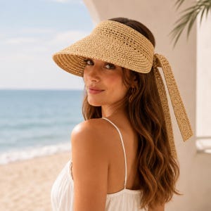 May include: A beige straw visor featuring a wide, curved brim and a long, matching woven ribbon tie at the back. The visor is constructed from a textured, braided material in a natural light tan color, designed to provide shade with an open top.