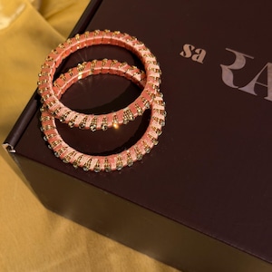 May include: Two circular bangles wrapped in peach-colored thread, featuring evenly spaced gold-toned metal accents and small clear crystals embedded along the outer circumference, resting on a dark brown box with partial white text visible.