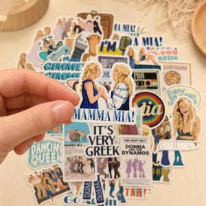 Könnte beinhalten: Eine Sammlung von Mamma Mia-Aufklebern mit verschiedenen Illustrationen, darunter ein zentraler Aufkleber, der zwei blonde Figuren zeigt, die Händchen halten, mit dem Text Mamma Mia sowie weitere Designs mit Text wie Dancing Queen.