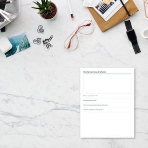 May include: A white paper sheet with the printed heading Monthly Brain Dump & Reflection, featuring a large blank rectangular box and four lined prompts for notes, positioned on a white marble surface with various office supplies nearby.