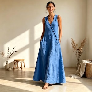 Puede incluir: Un vestido azul sin mangas que llega hasta el suelo, con escote en V y una fila diagonal de botones oscuros que se extiende desde el hombro hasta la cintura. La prenda tiene una silueta relajada y fluida con falda amplia y bolsillos.