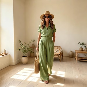 May include: A sage green short-sleeved button-down shirt and matching high-waisted wide-leg trousers. The outfit is paired with a straw fedora hat, dark sunglasses, and a woven straw tote bag. The clothing features a textured fabric finish.