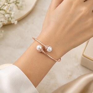 Può includere: Un bracciale aperto in oro rosa con due perle bianche sferiche alle estremità. Ogni perla è incastonata in una piccola coppetta metallica decorativa ornata da minuscole pietre chiare e scintillanti. Finitura liscia e lucida.