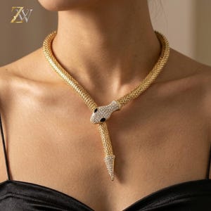 Puede incluir: Un collar de serpiente en tono dorado con una cadena de malla texturizada que rodea el cuello. La cabeza de la serpiente está incrustada con cristales claros y tiene dos ojos de piedra negra, con una cola adornada con cristales que cuelga.