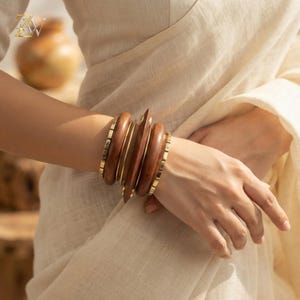 Puede incluir: Una pila de pulseras que consta de tres brazaletes de madera lisa de color marrón oscuro intercalados con dos brazaletes finos de metal dorado pulido con un diseño de eslabones rectangulares segmentados, sobre un fondo de tela color crema.