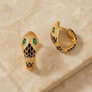 Peut inclure: Une paire de créoles dorées en forme de tête de serpent, ornées d'écailles en émail noir, de cristaux pavés transparents et de deux petites pierres vertes taille marquise en guise d'yeux. Les boucles ont une fermeture à charnière.