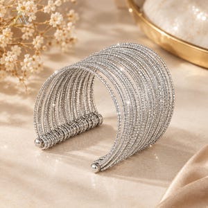 May include: A wide cuff bracelet composed of multiple thin, silver-toned wire bands arranged in a parallel formation. The surface of each wire is textured with a fine, reflective finish that catches the light. The bracelet features a C-shaped opening.