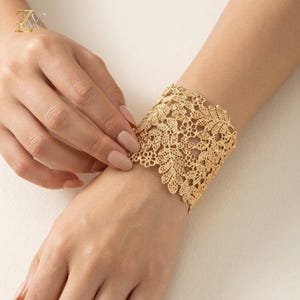 May include: A wide gold cuff bracelet featuring an intricate, openwork lace pattern of delicate flowers and leaves. The metallic gold finish has a textured, filigree-style design that wraps around the wrist, creating a detailed botanical motif.