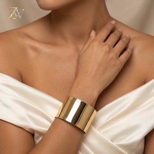 Könnte beinhalten: Ein breites, poliertes goldfarbenes Manschettenarmband mit einer glatten, reflektierenden Oberfläche. Das Armband hat ein minimalistisches, offenes Design, das sich um das Handgelenk legt und ein glänzendes Metallfinish aufweist.