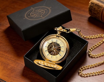 May include: A gold-tone mechanical pocket watch with a skeleton dial and Roman numerals, featuring a matching gold-tone chain with a belt clip. The watch is displayed inside an open black box with a compass graphic on the lid.