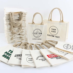 Peut inclure: Une collection de sacs fourre-tout en toile blanc cassé avec des anses beiges, présentant divers motifs imprimés en noir et rouge, incluant le texte CUSTOM LOGO HERE, YOUR LOGO CUSTOM TEXT, CUSTOM SECOND TEXT HERE et YOUR PHONE HERE, disposés en pile.