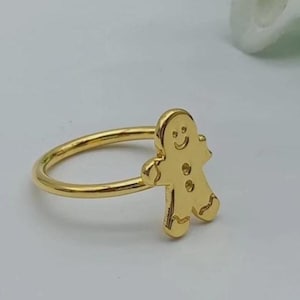 Puede incluir: Un anillo de tono dorado que presenta una figura plana y estilizada de hombre de jengibre unida a la banda. El hombre de jengibre tiene una cara sonriente grabada, dos botones circulares en el torso y líneas onduladas en las extremidades.