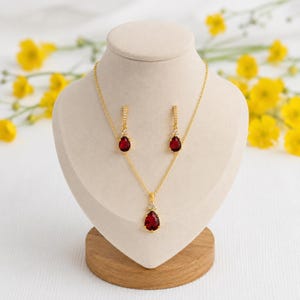 Puede incluir: Un conjunto de collar y pendientes de oro con piedras preciosas rojas en forma de lágrima. El colgante del collar y cada pendiente están acentuados con pequeñas piedras transparentes engastadas en oro sobre un expositor de color beige.
