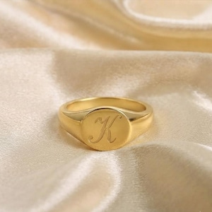 May include: A gold-toned signet ring featuring a smooth, polished surface with the letter K engraved in a cursive script on the circular face. The ring has a solid band that tapers slightly toward the back, showcasing a metallic finish.