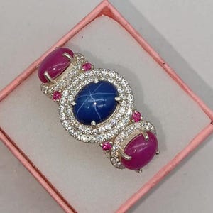 Op de afbeelding: Een zilveren ring met een centrale ovale blauwe stersaffier omringd door een halo van kleine heldere stenen. Twee ovale roze cabochonstenen zijn aan de uiteinden van de ring geplaatst, geaccentueerd door kleine roze steentjes in het zilver.