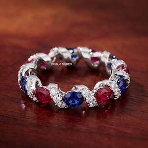 May include: A silver eternity band featuring alternating oval-cut red rubies and blue sapphires, interspersed with small round white diamonds set in a twisted metal design. The text House Of Kevadiya is visible on the inner band surface.