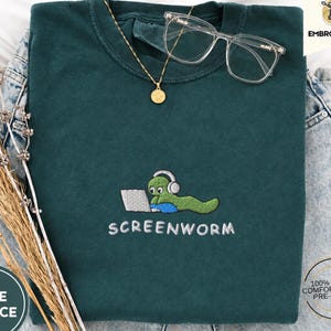 Könnte beinhalten: Ein dunkelgrünes T-Shirt mit Rundhalsausschnitt und einer Stickerei, die einen grünen Wurm mit weißen Kopfhörern an einem silbernen Laptop zeigt. Der Text "SCREENWORM" ist darunter in Weiß gestickt. Das T-Shirt besteht aus 100% Baumwolle.