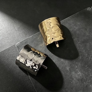 May include: Two wide cuff bracelets, one in a polished gold tone and one in a polished silver tone. Both bracelets feature a distinct hammered, irregular surface texture that creates a rippled, reflective appearance across the curved metal bands.