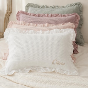 Könnte beinhalten: Ein Stapel aus vier rechteckigen Kissen mit Rüschenkanten und Waffelstrickstruktur. Die Kissen sind in Weiß, Hellrosa, Altrosa und Salbeigrün gehalten. Auf dem weißen Kissen ist der Name Olivia in einer kursiven Schreibschrift eingestickt.