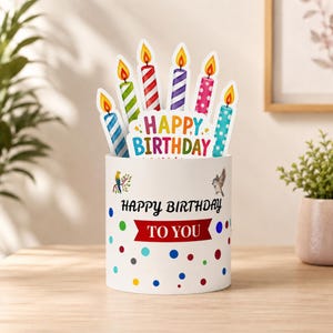 May include: A white cylindrical container decorated with colorful polka dots, a small bird illustration, and the text "HAPPY BIRTHDAY TO YOU". A pop-up card insert features six lit birthday candles and the text "HAPPY BIRTHDAY" in colorful letters.