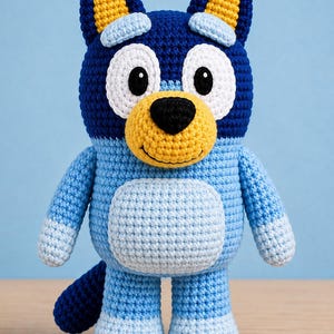 May include: A crochet plush toy of a blue dog character with large white eye patches, a yellow muzzle, a black nose, and dark blue ears with yellow inner sections. The body is light blue with a lighter blue belly patch and dark blue tail.