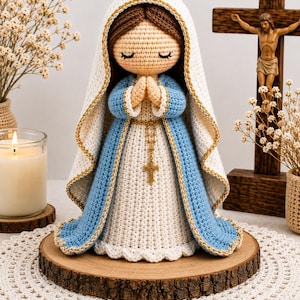 Puede incluir: Una muñeca de ganchillo de la Virgen María con los ojos cerrados y las manos en oración. La figura viste una túnica blanca y un manto azul con borde dorado, tiene cabello castaño y un pequeño rosario dorado colgado en la cintura.
