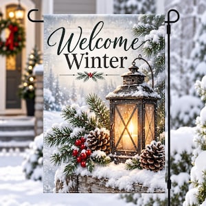 May include: A rectangular garden flag featuring the text Welcome Winter in black script and serif font. The image displays a dark metal lantern with a glowing candle, surrounded by snow-covered pine branches, red berries, and pinecones.