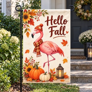 May include: A rectangular garden flag featuring a pink flamingo wearing an orange knit hat and a plaid scarf. The text Hello Fall is printed in brown. The design includes orange pumpkins, sunflowers, red berries, and autumn leaves on a cream background.