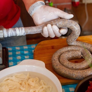 Puede incluir: Una máquina embutidora de metal extruye una salchicha de carne larga, marrón y enrollada en una tripa natural. Un cuenco blanco lleno de agua y tripas blancas adicionales se encuentra cerca sobre una superficie de cuadros azules y blancos.
