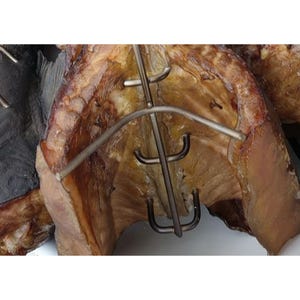 May include: A smoked fish fillet secured by a stainless steel metal rack. The fish has a golden-brown, textured surface with visible skin and flesh. The metal rack features a central vertical rod with curved prongs holding the fish in place.