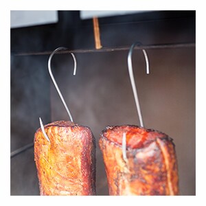 Könnte beinhalten: Zwei Stücke geräuchertes Fleisch hängen an silbernen S-Haken aus Metall. Das Fleisch hat eine gebräunte, strukturierte Oberfläche und ist an einer horizontalen Metallstange befestigt. Die Haken sind oben gebogen und durchstechen das Fleisch.