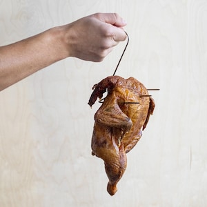 Könnte beinhalten: Ein ganzes gebratenes, goldbraunes Huhn, das an einem dünnen Metallhaken hängt. Das gegarte Geflügel hat eine knusprige, gewürzte Haut und wird von zwei horizontalen Metallspießen durchbohrt, die seine Form beim vertikalen Hängen stützen.