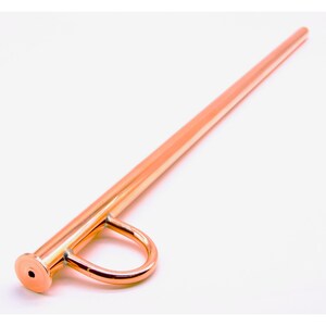 May include: A polished copper-colored metal stirring rod featuring a long, slender cylindrical shaft and a circular ring handle attached near the flat, circular end cap. The surface has a reflective, metallic finish throughout the entire object.