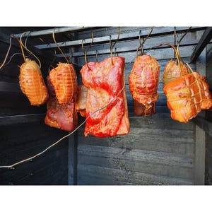 Könnte beinhalten: Mehrere Stücke geräuchertes Fleisch, darunter große Scheiben und runde Braten, hängen an Metallhaken in einem dunklen Räucherofen aus Holz. Das Fleisch hat eine glänzende, rotbraune Oberfläche und ist teilweise mit weißem Netzgarn umwickelt.