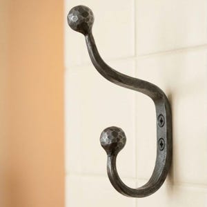 Puede incluir: Un gancho de pared de metal forjado a mano en color gris oscuro, con un diseño de doble gancho y un acabado martillado y texturizado. El gancho tiene dos extremos redondeados en forma de bola y se fija con dos tornillos negros visibles.