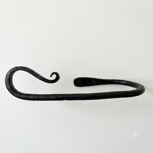 May include: A black hand-forged metal towel ring with a textured finish. The design features a curved, scroll-like end that loops back towards the mounting plate, which is secured by two visible screws on the flat, rounded attachment point.