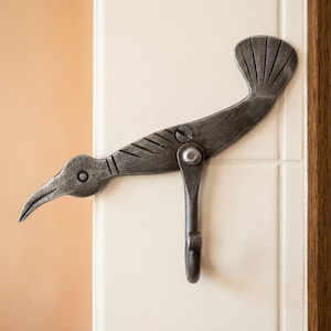 May include: A dark grey metal wall hook shaped like a bird with a long beak and a fanned tail. The bird body features etched line details and is attached to a downward-curving hook mechanism secured by a central metal screw and a pivot rivet.