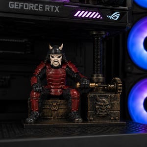 May include: A figurine of a samurai wearing red and black armor with a horned helmet, sitting on a bronze-colored chest. Next to the figure is a mechanical device with a gear and a bronze box featuring a sculpted demon face mask design.