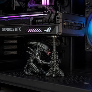 May include: A black Xenomorph figure from the Alien franchise acting as a GPU support bracket. The figure is positioned beneath a black GeForce RTX graphics card with purple lighting, holding up a metallic threaded rod that stabilizes the hardware.