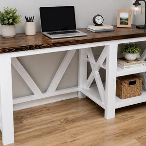 May include: A rectangular desk with a dark brown stained wood top and a white painted frame featuring X-shaped side supports. The desk includes an integrated side shelving unit with two open shelves, one containing a woven basket and books.