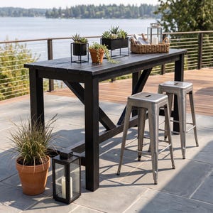 Peut inclure: Une table de bar haute en bois noir avec des supports latéraux en forme de X, accompagnée de deux tabourets de bar en métal argenté. La table porte trois plantes succulentes, un panier tressé avec une lanterne, et une lanterne au sol.