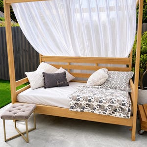 May include: A light wood canopy daybed with a white sheet, a patterned throw blanket, and several decorative pillows, including one dark gray rectangular pillow with the word home embroidered on it, topped with sheer white curtains.