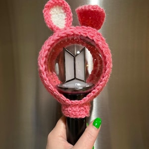 May include: A pink crocheted bunny ear cover for a light stick. The cover has white inner ears and fits over a clear, round light. The light stick has a black handle and a logo in the center.