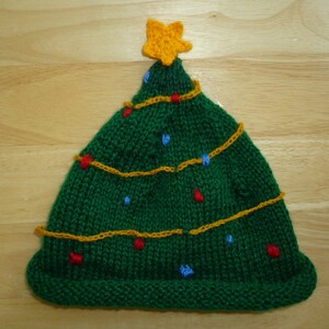 May include: A green knitted Christmas tree hat with a yellow star on top. The hat is decorated with yellow, red, and blue yarn to resemble Christmas lights.