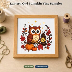 Op de afbeelding: Een kruissteekpatroon getiteld Lantern Owl Pumpkin Vine Sampler met een bruine uil op een tak met oranje pompoenen, rode bladeren en een verlichte lantaarn. Het ontwerp is ingelijst in licht hout en bevat labels voor PDF-patroon en zelfverzekerde beginner.
