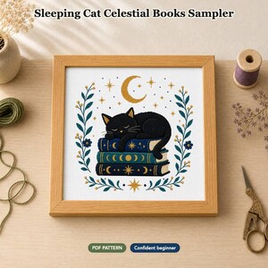 Op de afbeelding: Een kruissteekpatroon getiteld Sleeping Cat Celestial Books Sampler met een zwarte kat die slaapt op een stapel van drie blauwe boeken. Het ontwerp bevat een gouden halve maan, sterren en een bloemenkrans, ingelijst in lichtgekleurd hout.