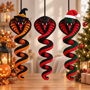 May include: Three hanging crochet cobra ornaments with coiled bodies. The left snake is black and orange with a witch hat, the center is black and red, and the right is black and green with a red and white Santa hat. All have red eyes and tongues.